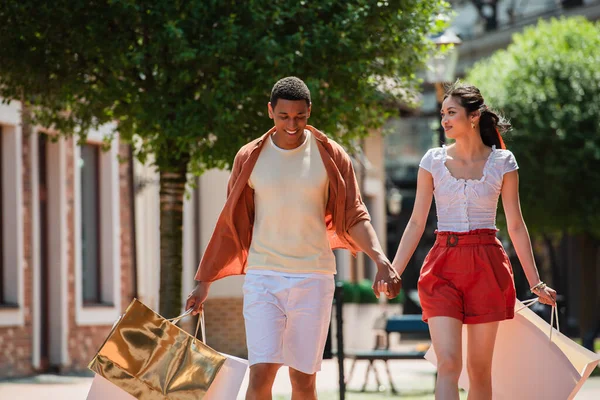 Young multiethnic couple in summer clothes walking with purchases on urban street — Stock Photo