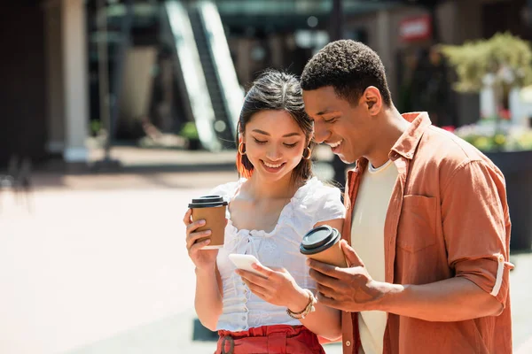 Alegre interracial casal com copos de papel olhando para celular ao ar livre — Fotografia de Stock