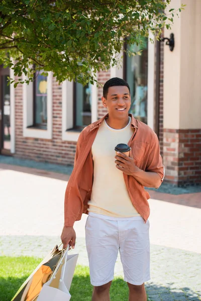 Glücklich afrikanisch-amerikanischer Mann mit Imbissgetränk zu Fuß mit Einkaufstüten in der Stadt — Stockfoto