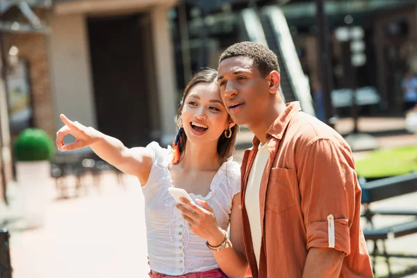 Amazed asian woman with smartphone pointing with finger near african american man — Stock Photo