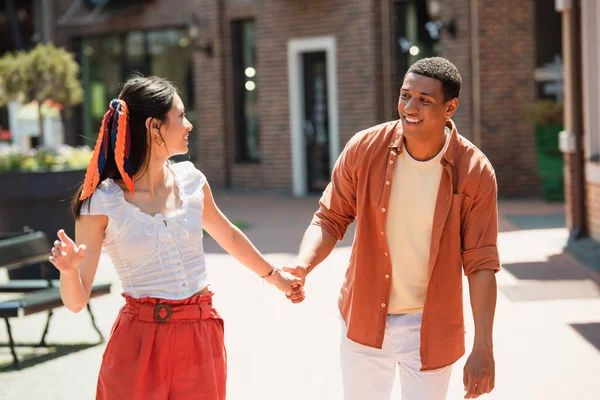 Cheerful interracial couple in summer clothes holding hands on city street — Stock Photo