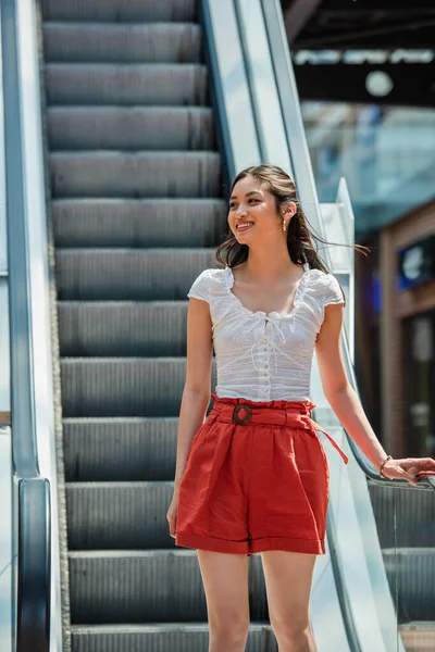 Feliz asiático mujer en verano ropa mirando lejos en escaleras mecánicas - foto de stock