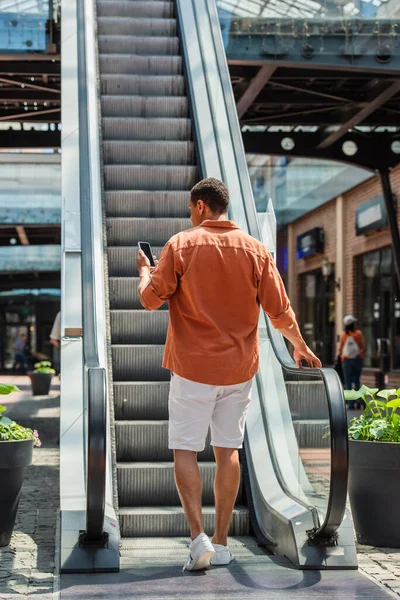 Vista posterior del hombre afroamericano utilizando el teléfono móvil en la escalera mecánica - foto de stock