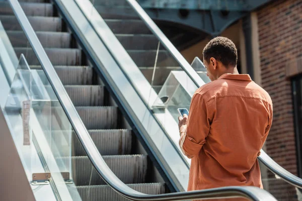 Jeune homme afro-américain en chemise de messagerie sur téléphone mobile sur escalator — Photo de stock