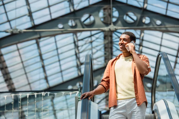 Усміхнений афроамериканський чоловік дивиться геть, розмовляючи на смартфоні на ескалаторі — стокове фото
