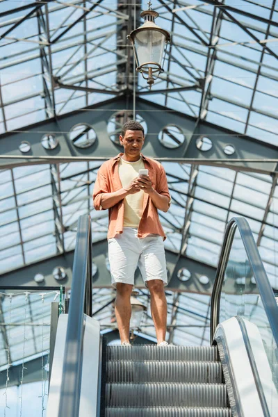 Vista de ángulo bajo del hombre afroamericano utilizando el teléfono celular en la escalera mecánica - foto de stock