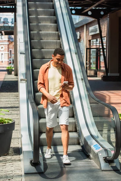 Homem americano africano alegre em shorts usando telefone celular perto escada rolante — Fotografia de Stock