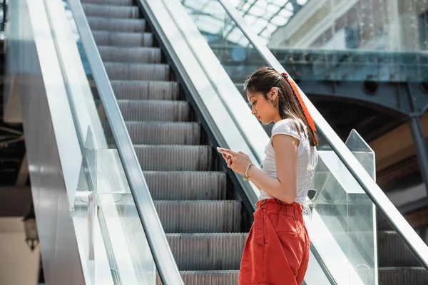 Joven asiático mujer mensajería en móvil teléfono en escaleras mecánicas - foto de stock