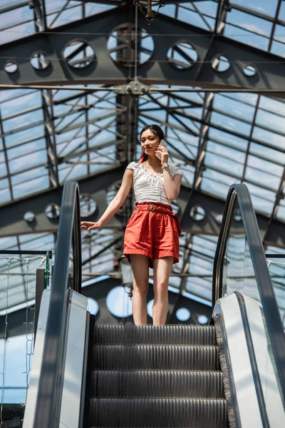 Bajo ángulo vista de elegante asiático mujer hablando en celular en escalera mecánica - foto de stock