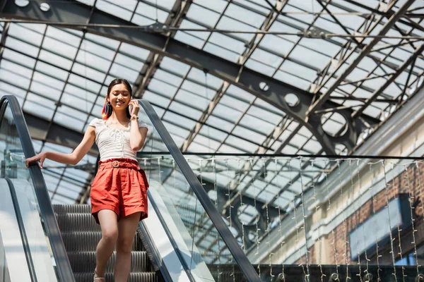 Low angle view of pleased asian woman talking on mobile phone on escalator — Stock Photo