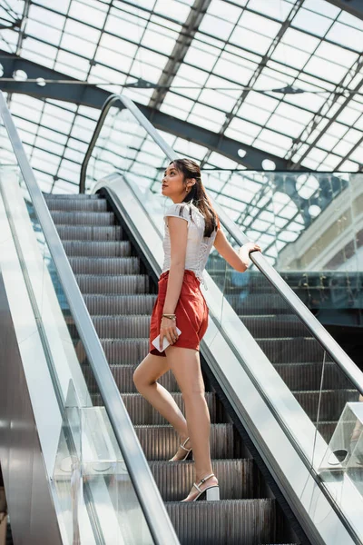 Joven asiático mujer holding smartphone mientras mirando lejos en escaleras mecánicas - foto de stock