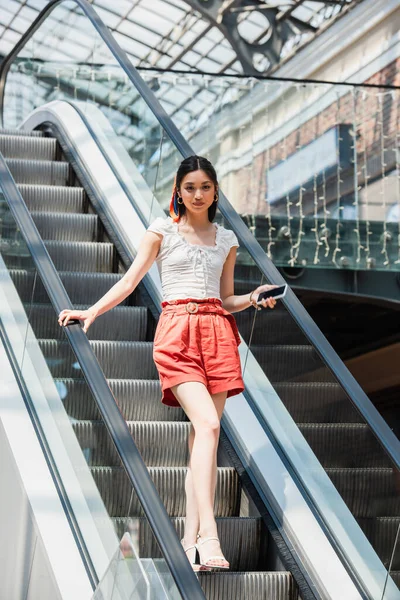 Young asian woman in shorts looking at camera while standing with smartphone on escalator — Stock Photo