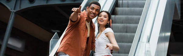 Cheerful african american man pointing with finger near asian woman on escalator, banner — Stock Photo