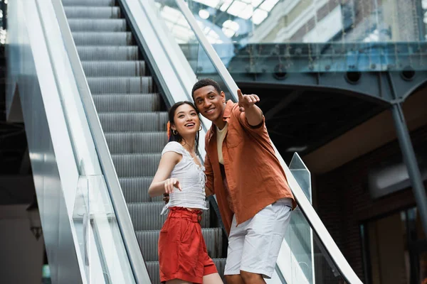 Excited asian woman looking away near african american man pointing with finger on escalator — Stock Photo