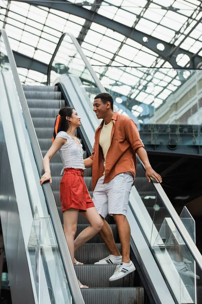 Feliz pareja multiétnica en ropa de verano mirándose mutuamente en escaleras mecánicas - foto de stock