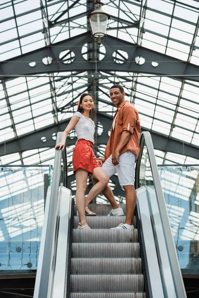 Low angle view of interracial couple in stylish summer clothes smiling at camera on escalator — Stock Photo