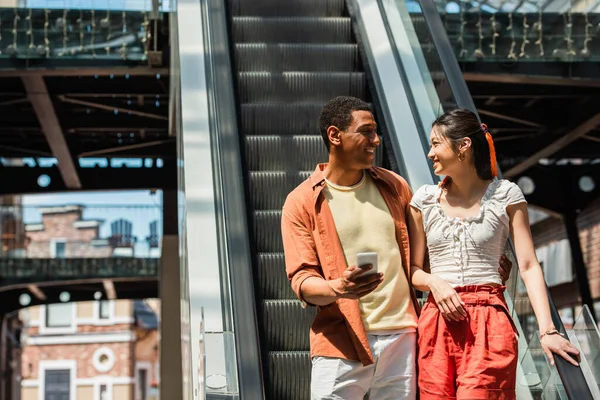 Elegante africano americano hombre con smartphone sonriendo a asiático novia cerca de escaleras mecánicas - foto de stock