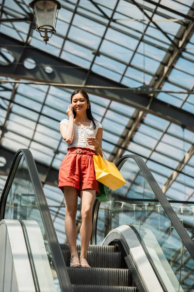 Alegre asiático mujer con compras hablando en smartphone en escalera mecánica - foto de stock