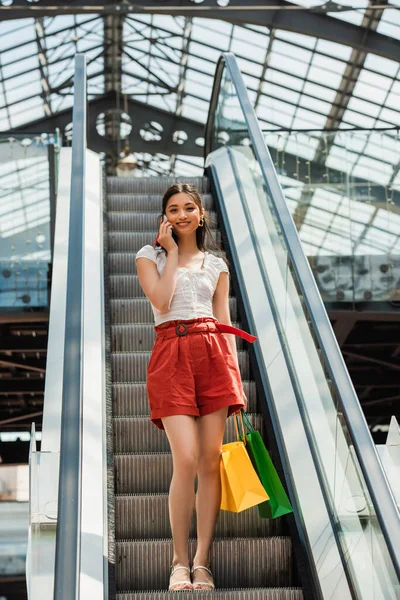 Alegre asiático mujer con compras hablando en celular en escalera mecánica - foto de stock