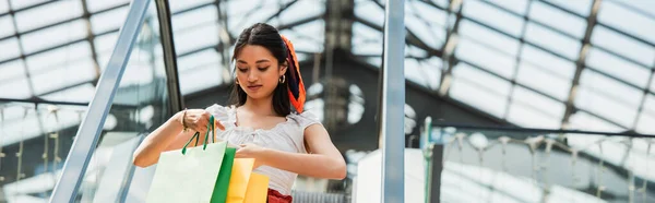 Stylische asiatische Frau schaut in Einkaufstüten auf Rolltreppe, Banner — Stockfoto