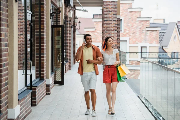 Smiling african american man calling on mobile phone while walking with asian woman — Stock Photo