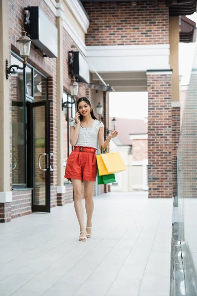 Volle Länge Ansicht der stilvollen asiatischen Frau zu Fuß mit Einkaufstaschen, während Gespräche auf dem Smartphone — Stockfoto