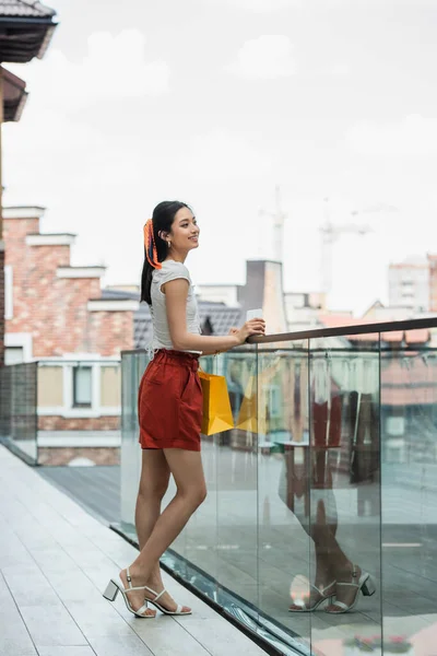 Feliz asiático mujer en verano ropa mirando lejos cerca de vidrio valla en ciudad - foto de stock