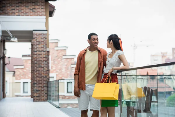Elegante mujer asiática con bolsa de compras mirando sonriente africano americano hombre al aire libre - foto de stock