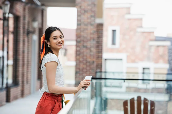 Cheerful asian woman with mobile phone looking at camera near fence in city — Stock Photo