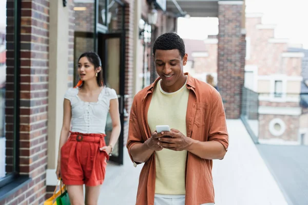 Alegre africano americano hombre usando móvil cerca asiático novia con compras bolsas - foto de stock