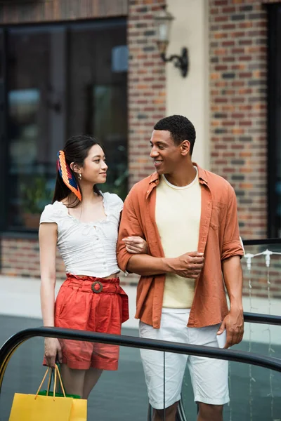 Stylish asian woman with shopping bag smiling at african american man outdoors — Stock Photo