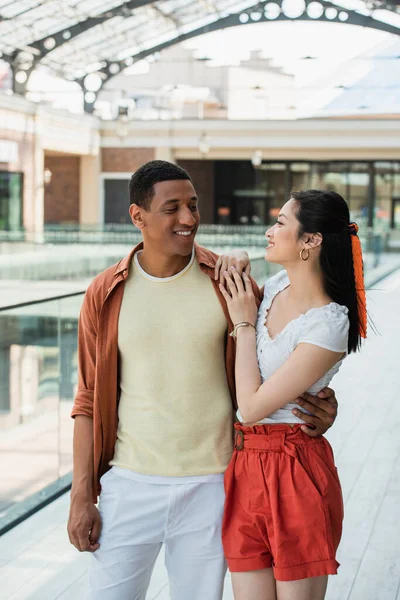 Stylish multiethnic couple looking at each other during summer walk in city — Stock Photo