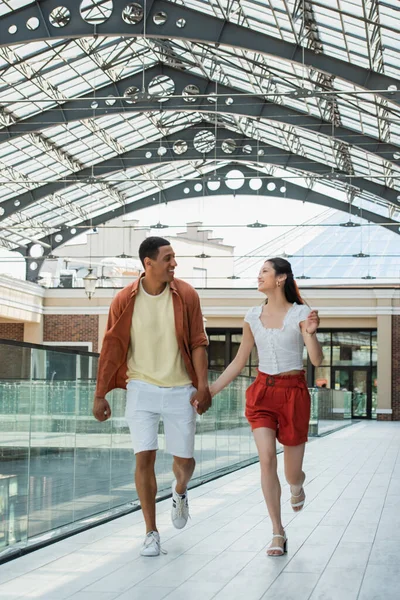 Excited multiethnic couple holding hands while running under glass roof in city — Stock Photo
