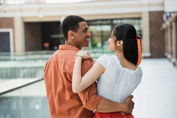 Heureux couple interracial dans des vêtements élégants embrasser et regarder l'autre à l'extérieur — Photo de stock