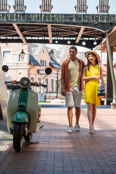Stylish multiethnic couple looking at scooter while walking on urban street — Stock Photo
