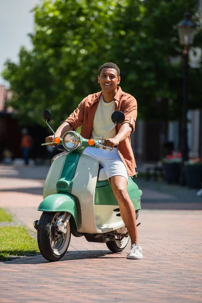 Joven afroamericano hombre en scooter sonriendo a cámara en la calle urbana — Stock Photo