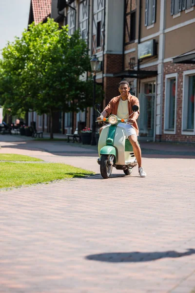 Hombre afroamericano satisfecho en scooter cerca de edificios en la calle urbana - foto de stock