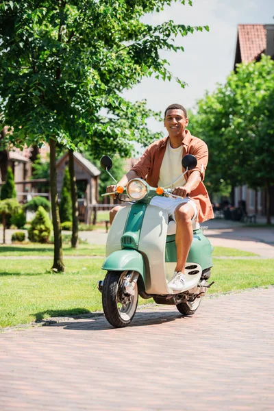 Hombre afroamericano positivo mirando a la cámara mientras monta scooter en la calle verde - foto de stock