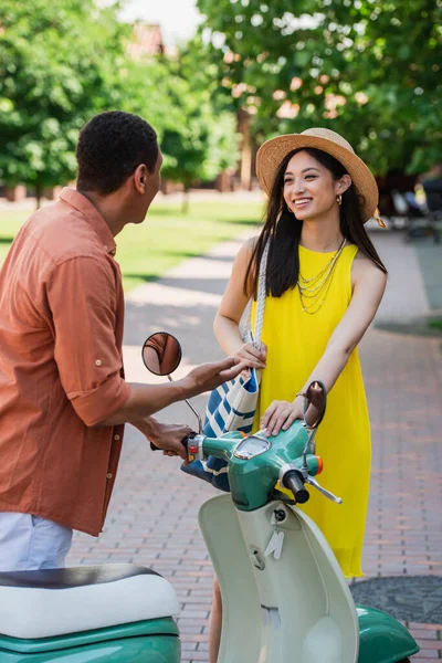 Africano americano hombre hablando a elegante asiático mujer cerca scooter al aire libre - foto de stock