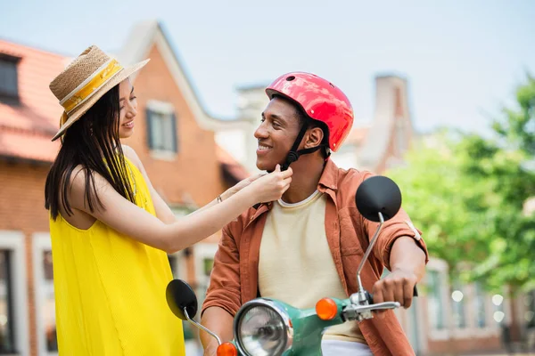 Asian woman in straw hat fastening helmet of african american man on scooter — Stock Photo