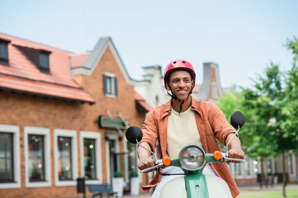 Sonriente afroamericano hombre en casco a caballo scooter en la ciudad - foto de stock