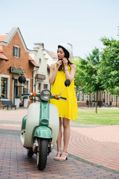Jeune femme asiatique en jaune sundress fixation casque près de scooter à l'extérieur — Photo de stock