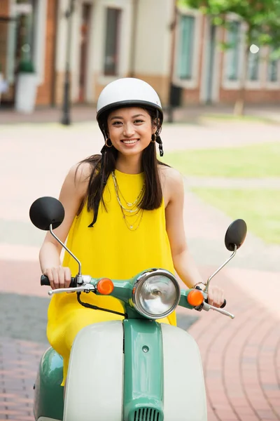 Joyful asian woman in hardhat and yellow sundress riding scooter in city — Stock Photo