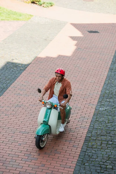 Vista ad alto angolo di uomo africano americano in hardhat cavalcare scooter sul marciapiede — Foto stock