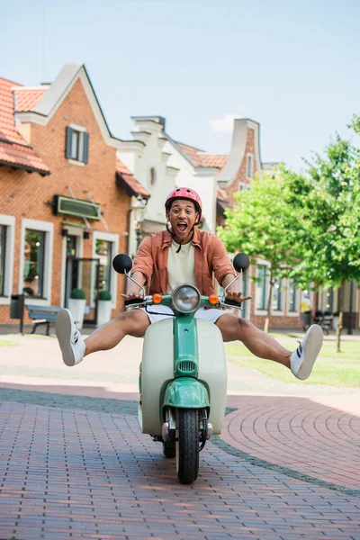 Emocionado hombre afroamericano levantando piernas mientras monta scooter al aire libre — Stock Photo