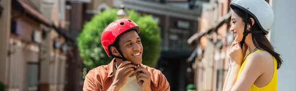 Fröhliches gemischtrassiges Paar, das draußen Helme befestigt, Banner — Stockfoto