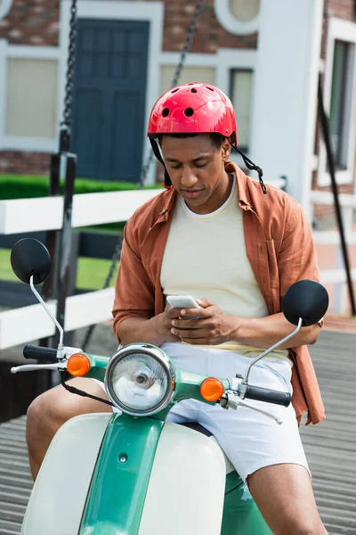 African american man in helmet messaging on smartphone while sitting on scooter — Stock Photo