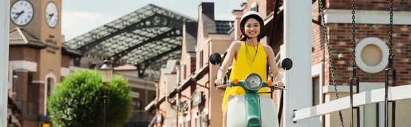 Heureux asiatique femme dans hardhat appréciant équitation scooter sur urbain rue, bannière — Photo de stock