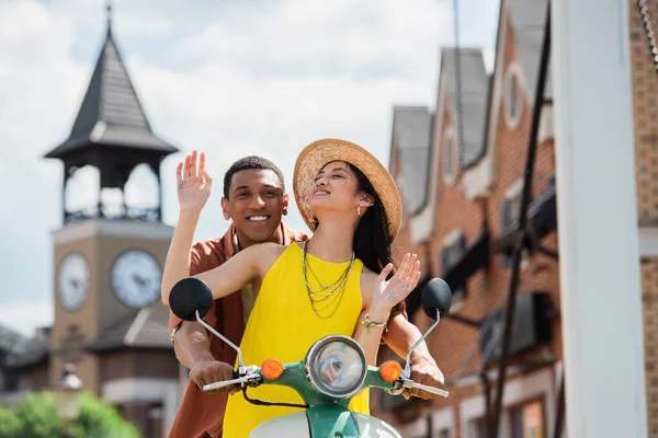 Alegre asiático mujer en paja sombrero a caballo scooter con feliz africano americano hombre - foto de stock