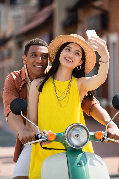 Alegre asiático mujer tomando selfie con africano americano novio en scooter - foto de stock